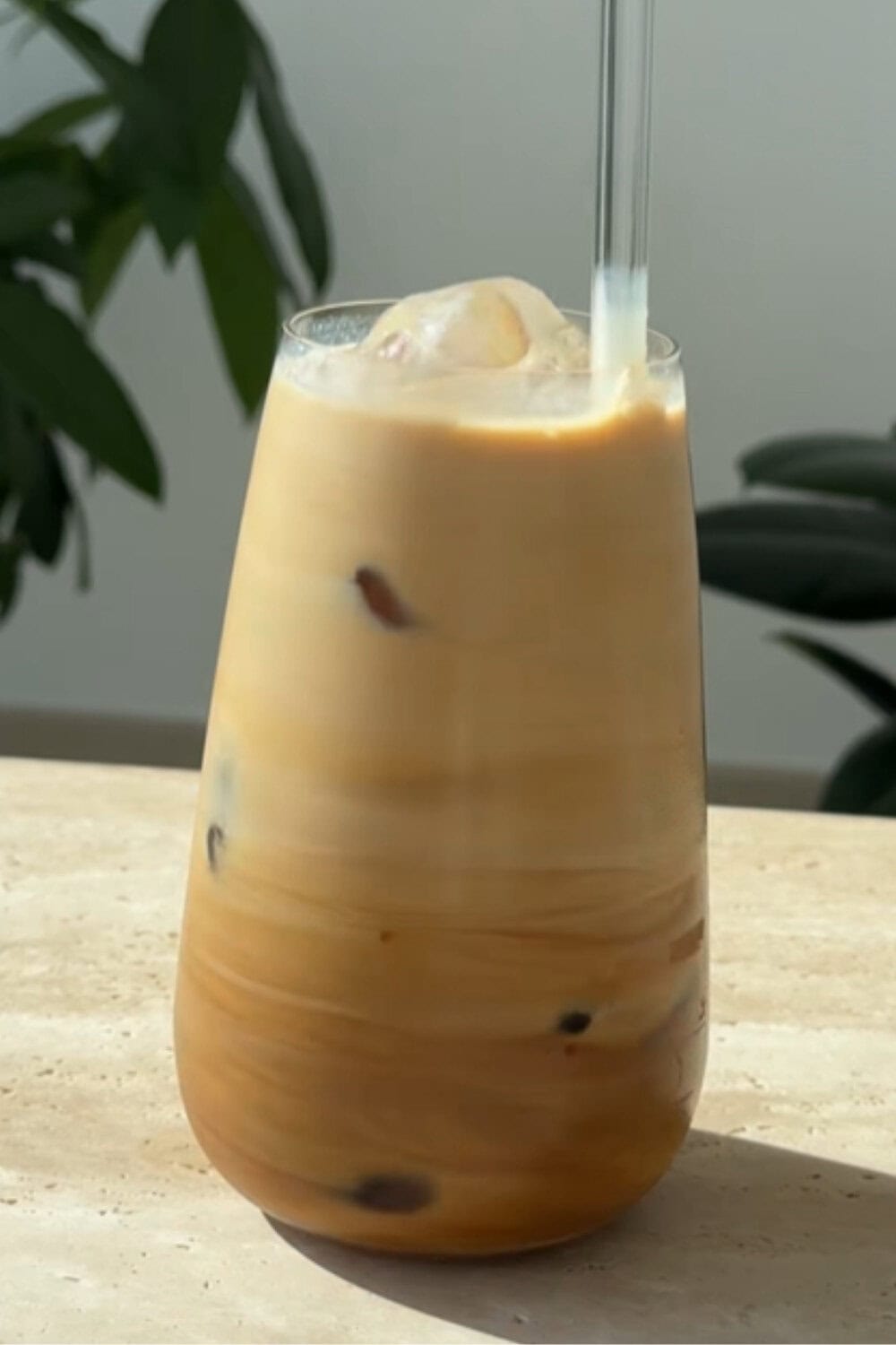 A tall glass of creamy iced coffee sitting on a sunlit countertop, with visible swirls of coffee and milk blending together and a clear straw on top, illustrating a simple iced coffee recipe.