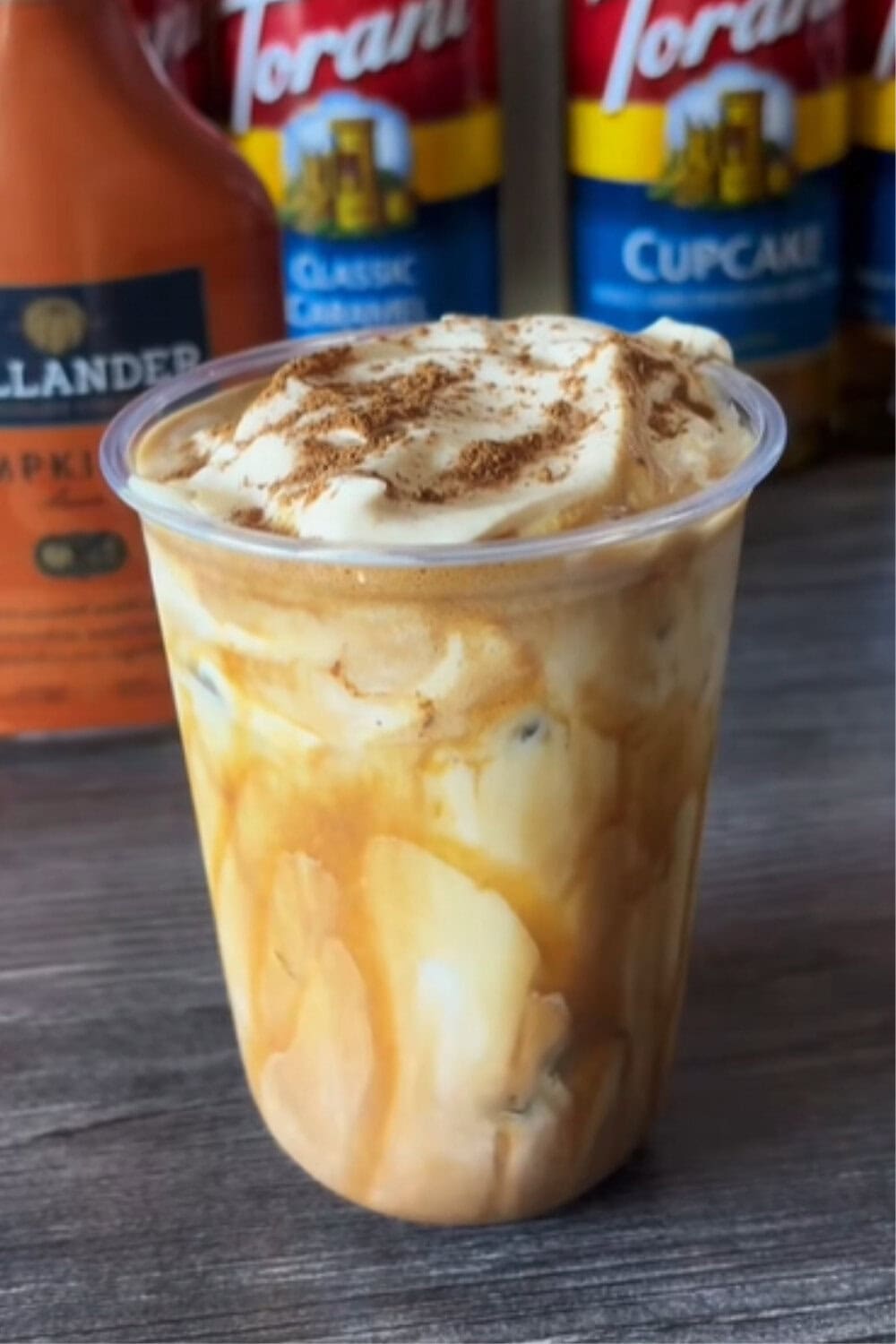 Iced coffee recipe made with pumpkin and white chocolate, served in a plastic cup with creamy swirls, whipped topping, and a sprinkle of cinnamon.