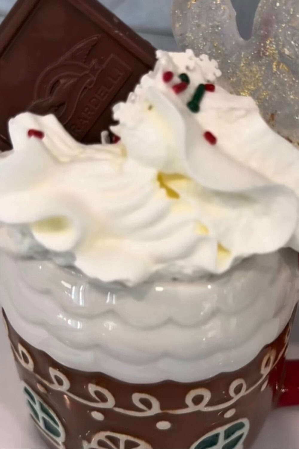 Iced coffee recipe topped with whipped cream, festive sprinkles, and a square of chocolate in a gingerbread-style holiday mug.