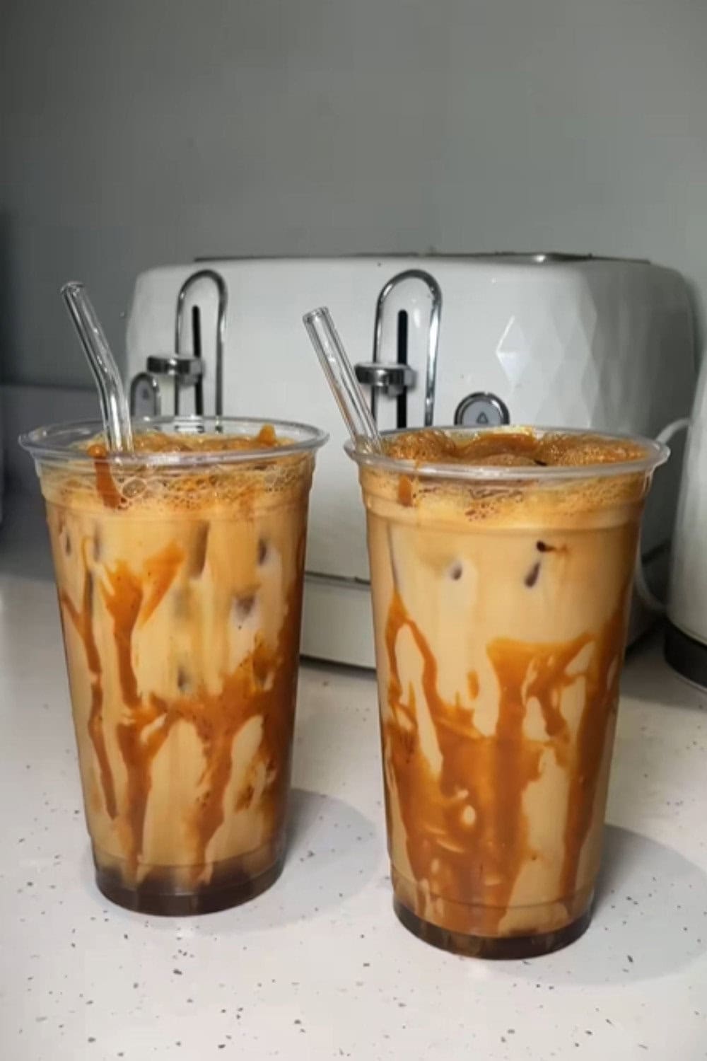 Two plastic cups filled with caramel-drizzled iced coffee topped with foam, each with a clear straw, set on a kitchen counter in front of a toaster, showing a caramel iced coffee recipe.