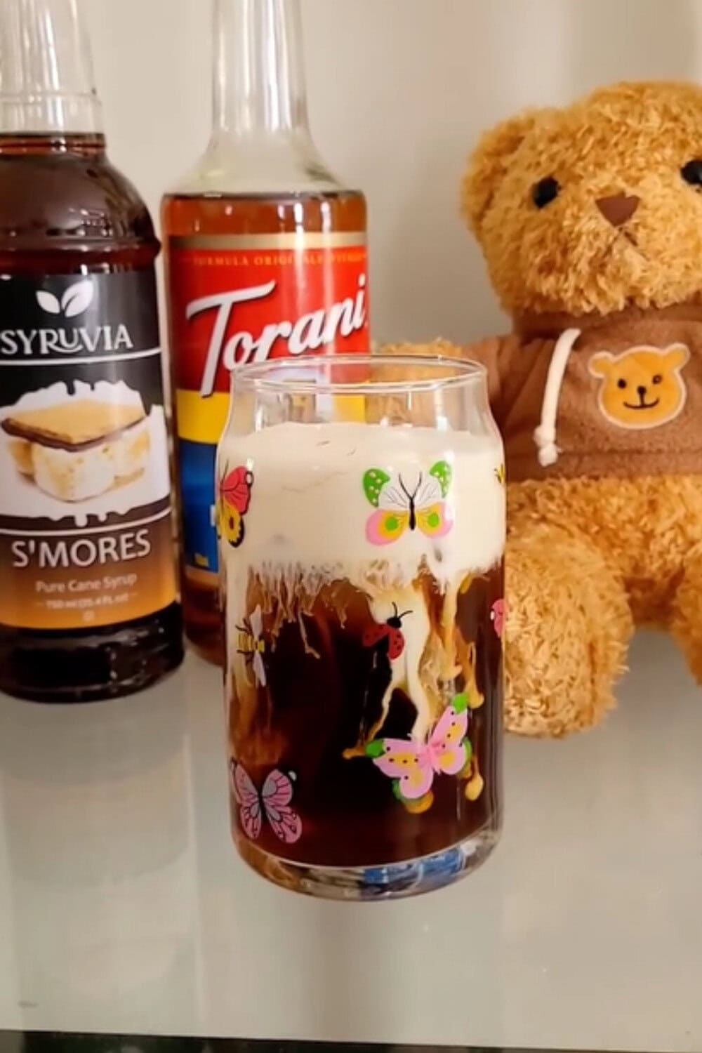 Iced coffee recipe with s&rsquo;mores-inspired flavor, topped with creamy foam and served in a butterfly-decorated glass beside syrup bottles.