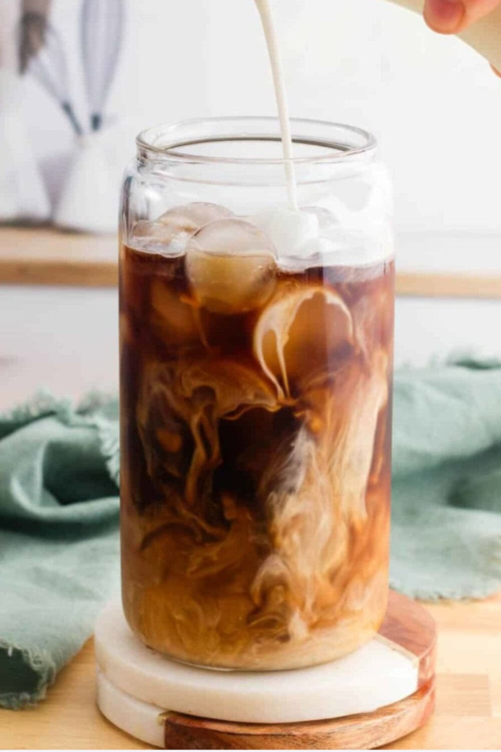 A glass jar filled with dark iced coffee as vanilla sweet cream is poured in, creating swirling patterns, with a soft kitchen background, demonstrating a vanilla sweet cream iced coffee recipe.