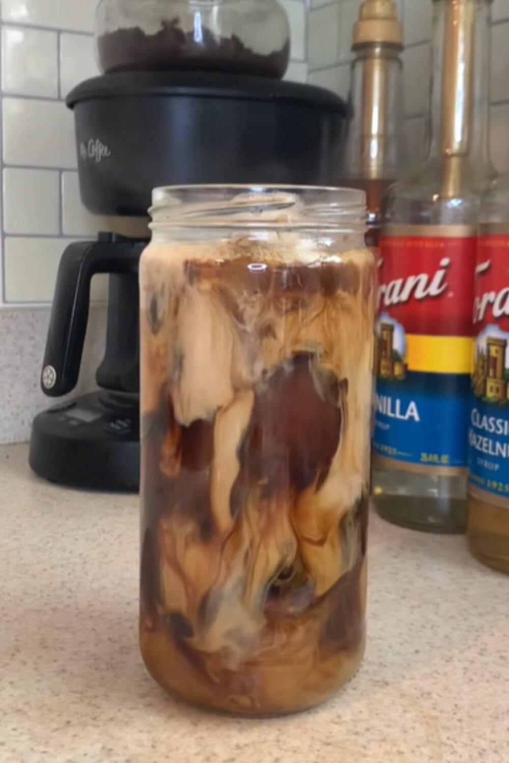 A jar-shaped glass filled with iced coffee and dramatic milk swirls, sitting on a kitchen counter near coffee syrups and a coffee maker, representing a protein iced coffee recipe.