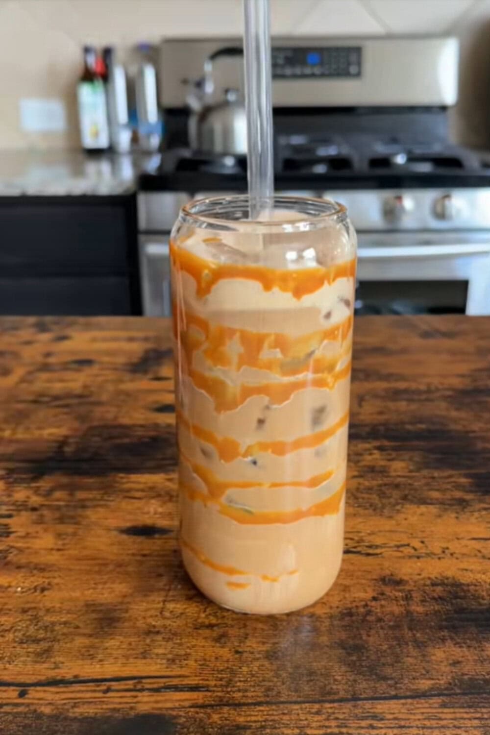 Iced coffee recipe featuring a can-shaped glass drizzled with thick dulce de leche and filled with creamy iced coffee, sitting on a rustic wooden table in front of a kitchen stove.