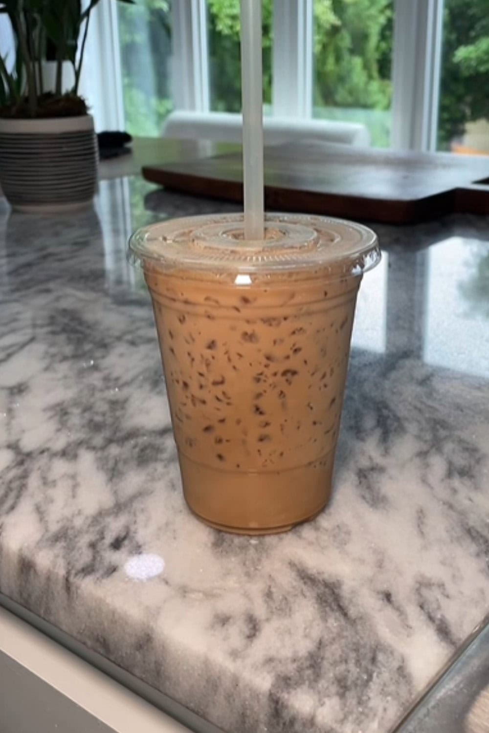 Iced coffee recipe showing a plastic cup packed with tiny ice pieces and creamy brown sugar coffee, placed on a marble counter with bright windows in the background.