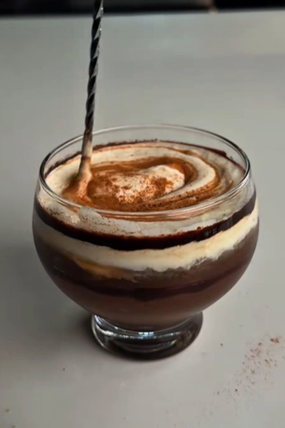 Iced coffee recipe with rich chocolate layers, swirls of cream, and a dusting of cocoa in a round glass cup.